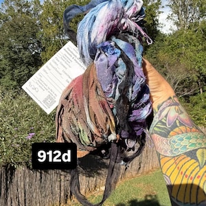 May include: A bundle of hand-dyed, textured fabric strips in shades of blue, purple, brown, and black. The bundle includes a white product tag with the text 912d, fiber content details, and care instructions, all held together in a loose, frayed arrangement.