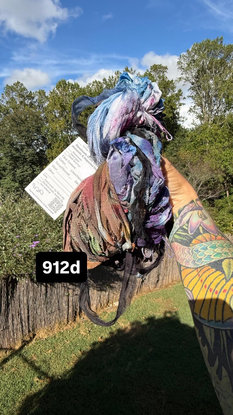 May include: A bundle of hand-dyed, textured fabric strips in shades of blue, purple, brown, and black. The bundle includes a white product tag with the text 912d, fiber content details, and care instructions, all held together in a loose, frayed arrangement.