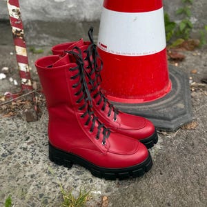 May include: A pair of red leather combat boots with black laces and a chunky black sole. The boots are mid-calf height and have a classic lace-up design. The boots are set against a background with a red and white traffic cone.