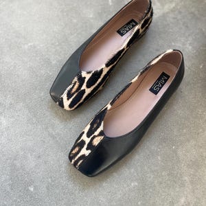 May include: A pair of black leather flats with a leopard print detail on the toe. The shoes have a square toe and a low heel. The brand name "MIAS" is printed on the inside of the shoes.