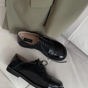 Op de afbeelding: Een paar zwarte leren veterschoenen met een laklederen neus. De schoenen hebben een ronde neus en een lage hak. De merknaam "MIAS" is zichtbaar aan de zijkant van een schoen.