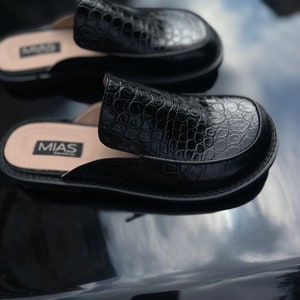 May include: A pair of black leather slip-on shoes with a crocodile pattern. The shoes have a rounded toe and a low heel. The brand name "MIAS" is printed on the insole.