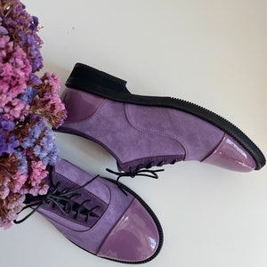 May include: A pair of purple suede lace-up shoes with a patent leather toe cap. The shoes have black laces and a black rubber sole.