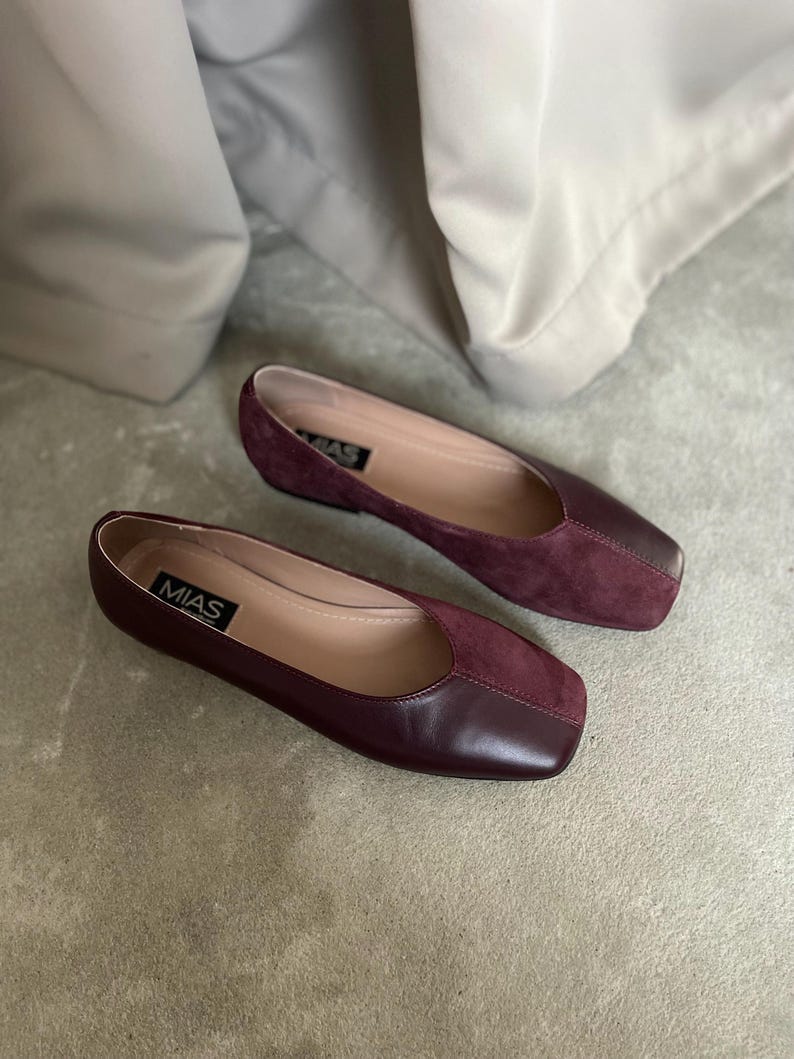 May include: A pair of burgundy leather flats with a square toe. The shoes have a suede panel on the top and the brand name "MIAS" is printed on the insole.