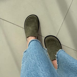 May include: A pair of olive green suede loafers with black soles. The loafers are worn with light blue denim jeans.