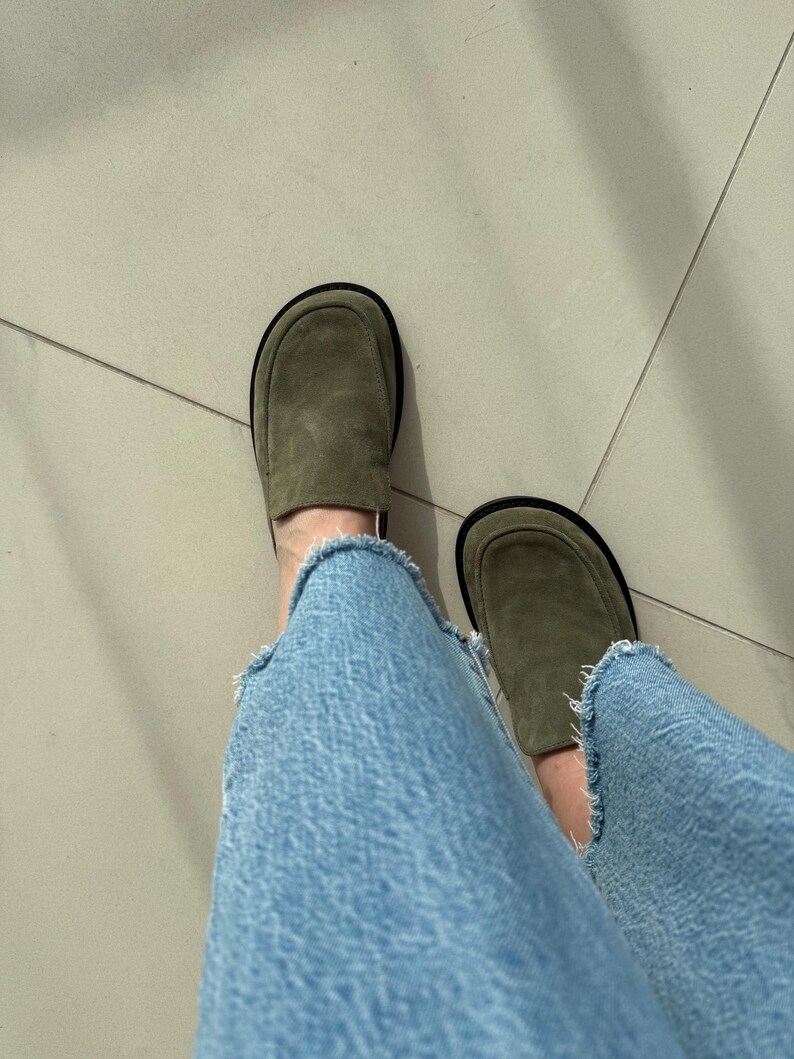 May include: A pair of olive green suede loafers with black soles. The loafers are worn with light blue denim jeans.
