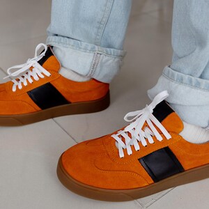 May include: A pair of orange suede sneakers with black and white accents. The sneakers have a gum sole and white laces.