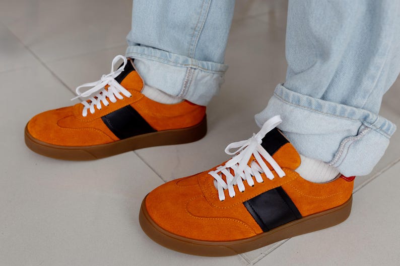 May include: A pair of orange suede sneakers with black and white accents. The sneakers have a gum sole and white laces.