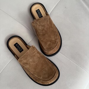 May include: A pair of brown suede slip-on shoes with black soles. The shoes have a simple design and the brand name "MIAS" is printed on the inside of each shoe.