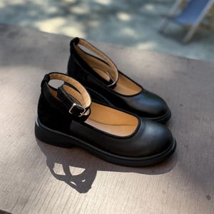 Könnte beinhalten: Ein Paar schwarze Mary Jane-Schuhe mit Knöchelriemen. Die Schuhe haben eine runde Spitze und einen kleinen Absatz. Die Riemen sind mit einer silbernen Schnalle befestigt. Die Schuhe sind aus Leder und Wildleder.