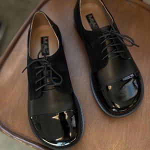 Op de afbeelding: Een paar zwarte leren veterschoenen met een laklederen neus. De schoenen staan op een houten stoel.