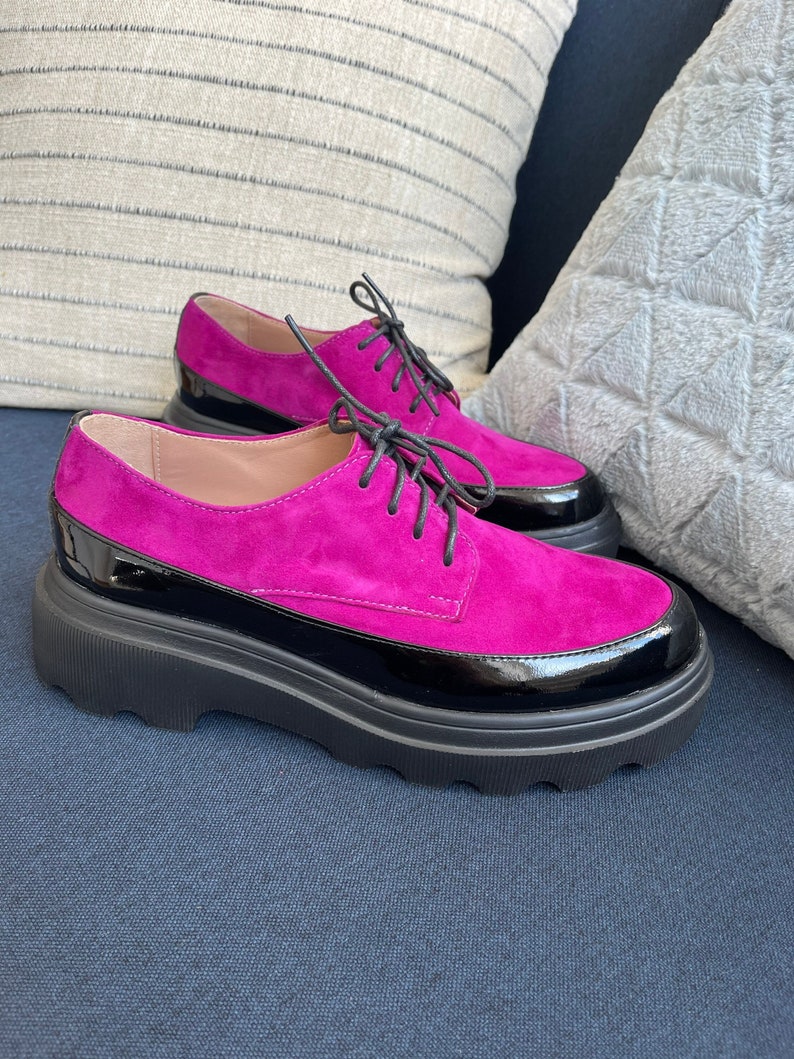 May include: A pair of pink suede shoes with black patent leather trim and chunky black soles. The shoes have lace-up closures.