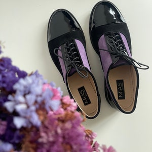 Könnte beinhalten: Ein Paar schwarze und lila Schnallenschuhe mit einer Lackspitze. Die Schuhe tragen den Markennamen "MIAS" auf der Innenseite.