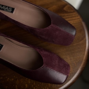 May include: A pair of burgundy flats with a square toe design. The shoes have a suede-like texture on the vamp and a smooth leather-like material on the toe cap. The brand name "MIAS" is visible on the side of one shoe.