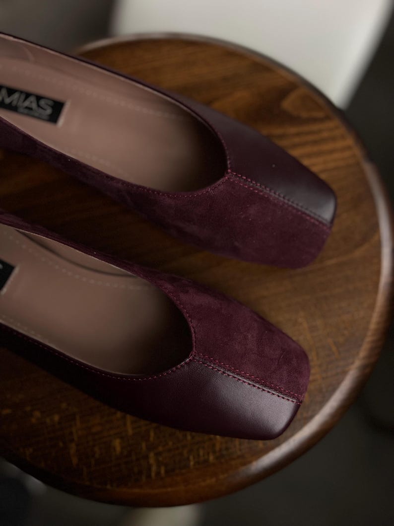 May include: A pair of burgundy flats with a square toe design. The shoes have a suede-like texture on the vamp and a smooth leather-like material on the toe cap. The brand name "MIAS" is visible on the side of one shoe.