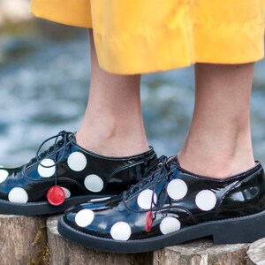 May include: Black patent leather lace-up shoes with white polka dots. The shoes have a rounded toe and a thick black sole.