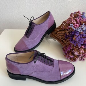 May include: A pair of purple suede and patent leather lace-up shoes with black soles. The shoes have a cap toe design and are sitting on a white surface.