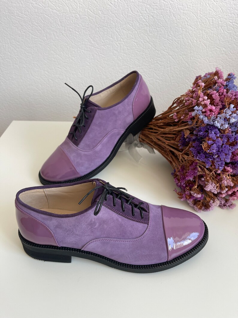 May include: A pair of purple suede and patent leather lace-up shoes with black soles. The shoes have a cap toe design and are sitting on a white surface.