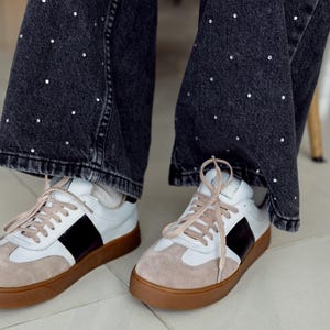 May include: A pair of white leather sneakers with black accents and brown soles. The sneakers have laces and are worn by a person wearing black jeans with white dots.