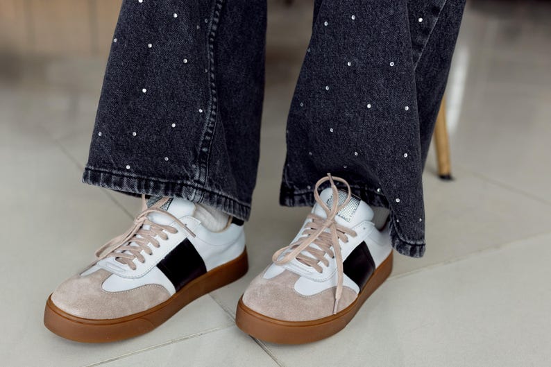 May include: A pair of white leather sneakers with black accents and brown soles. The sneakers have laces and are worn by a person wearing black jeans with white dots.
