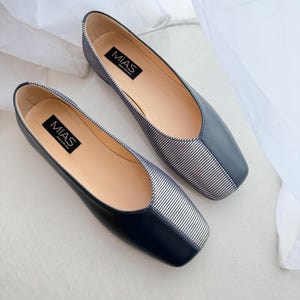 May include: A pair of black and white striped flats with a square toe. The shoes have a black leather upper and a white striped fabric toe. The brand name "MIAS" is printed on the insole of each shoe.