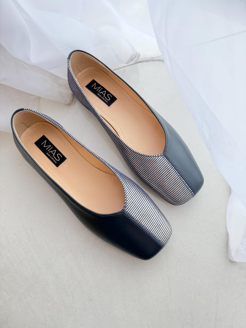 May include: A pair of black and white striped flats with a square toe. The shoes have a black leather upper and a white striped fabric toe. The brand name "MIAS" is printed on the insole of each shoe.