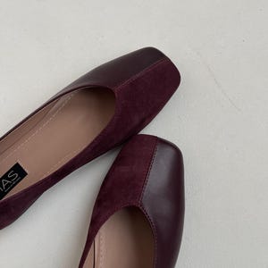 May include: A pair of burgundy suede and leather flats with a square toe. The shoes have a simple design and are made of high-quality materials.