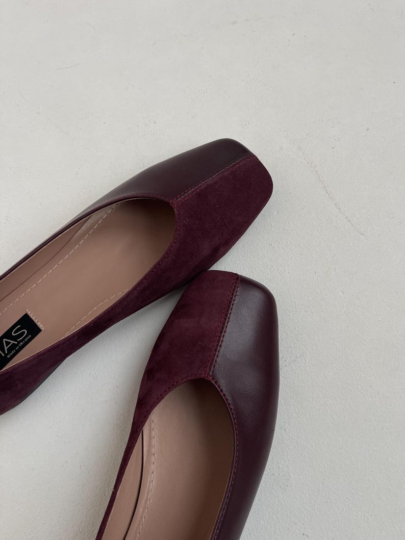 May include: A pair of burgundy suede and leather flats with a square toe. The shoes have a simple design and are made of high-quality materials.