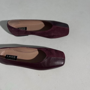 May include: Two pairs of burgundy flats with square toes. The shoes have a smooth leather upper and a contrasting suede vamp. The brand name "MIAS" is visible on the insole of one shoe. The shoes are designed for comfort and style.