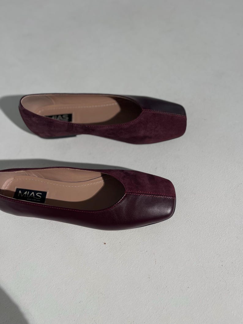 May include: Two pairs of burgundy flats with square toes. The shoes have a smooth leather upper and a contrasting suede vamp. The brand name "MIAS" is visible on the insole of one shoe. The shoes are designed for comfort and style.