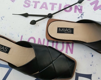 May include: A pair of black leather sandals with a woven design. The sandals have a square toe and a low heel. The sandals are on a white background with the words "Kensington Station London" printed on it.