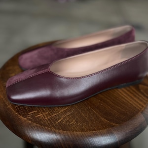May include: A pair of burgundy leather flats with a square toe. The shoes have a suede toe cap and a leather upper.