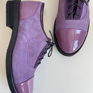 May include: A pair of purple suede and patent leather lace-up shoes with black laces. The shoes have a rounded toe and a black rubber sole.