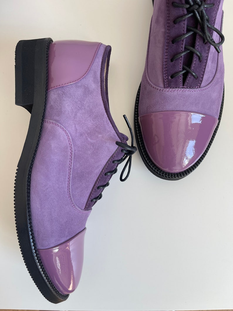 May include: A pair of purple suede and patent leather lace-up shoes with black laces. The shoes have a rounded toe and a black rubber sole.