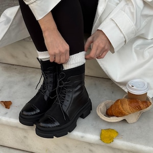 May include: Black lace-up combat boots with a chunky sole. The boots have a glossy toe cap and are paired with black leggings and white socks. A white coat and a croissant with coffee complete the look.