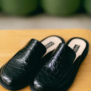 May include: A pair of black leather slip-on shoes with a crocodile-embossed pattern. The shoes have a rounded toe and a low heel.