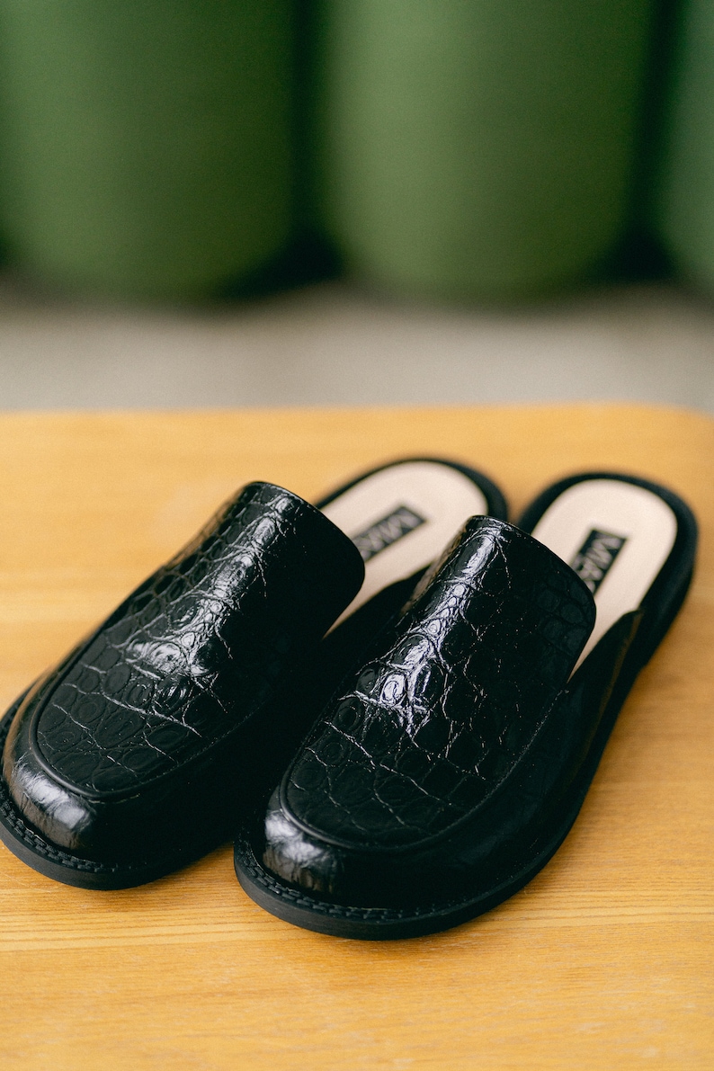 May include: A pair of black leather slip-on shoes with a crocodile-embossed pattern. The shoes have a rounded toe and a low heel.