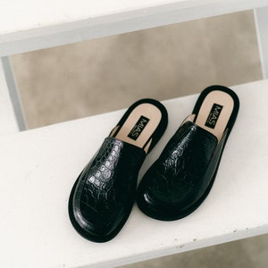 May include: A pair of black leather mules with a crocodile embossed pattern. The mules have a pointed toe and a low heel. The brand name "MIAS" is printed on the inside of each shoe.