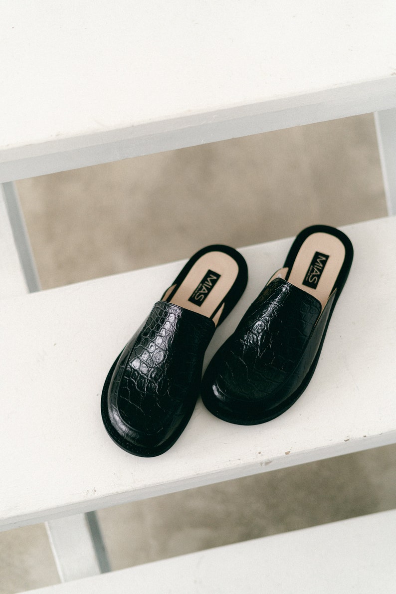May include: A pair of black leather mules with a crocodile embossed pattern. The mules have a pointed toe and a low heel. The brand name "MIAS" is printed on the inside of each shoe.