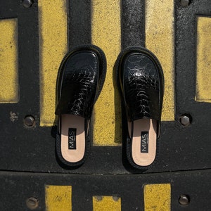 May include: A pair of black leather slip-on shoes with a crocodile-embossed pattern. The shoes have a black leather insole with the word "MIAS" printed on them.