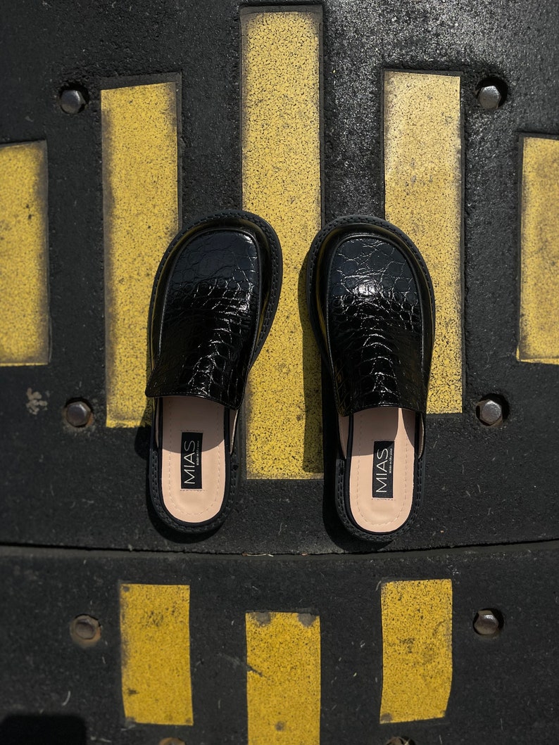 May include: A pair of black leather slip-on shoes with a crocodile-embossed pattern. The shoes have a black leather insole with the word "MIAS" printed on them.