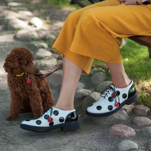May include: A woman wearing a yellow skirt and white shoes with black polka dots and red accents. A brown poodle is on a leash.