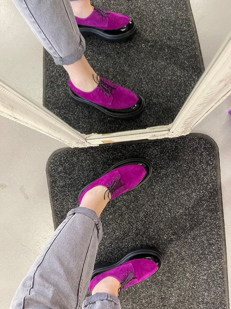 May include: A pair of purple suede shoes with black patent leather toes and black laces. The shoes have thick black soles.