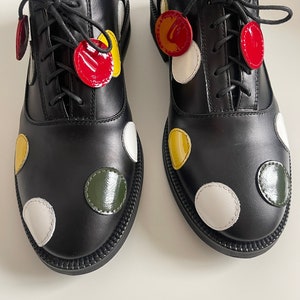May include: A pair of black leather oxford shoes with white, red, yellow, and green polka dots. The shoes have laces and are tied.