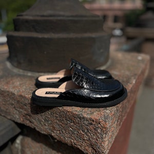 May include: Black leather slip-on shoes with a crocodile embossed pattern. The shoes have a rubber sole and the brand name "MIAS" is visible on the side.