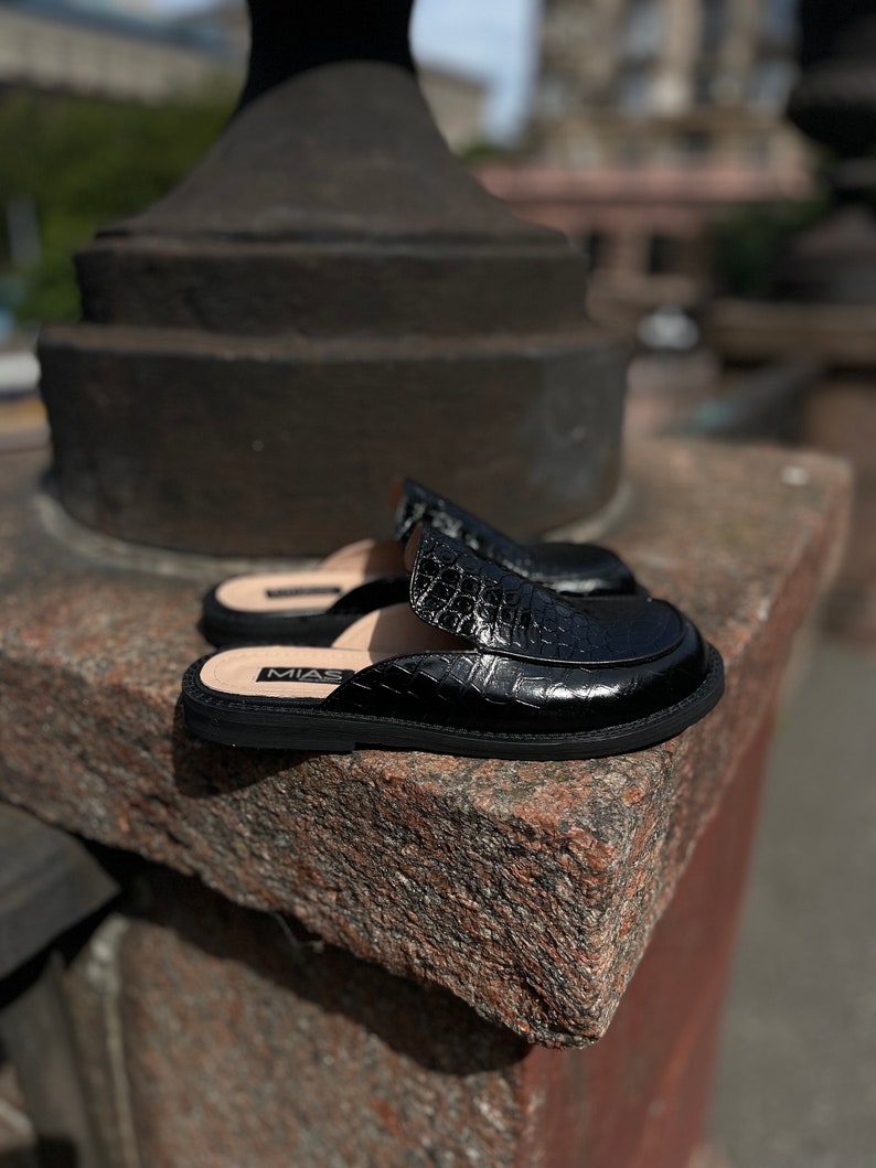 May include: Black leather slip-on shoes with a crocodile embossed pattern. The shoes have a rubber sole and the brand name "MIAS" is visible on the side.