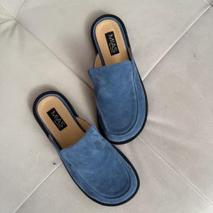 May include: A pair of blue suede slip-on shoes with black soles and the word "MIAS" printed on the inside of each shoe.