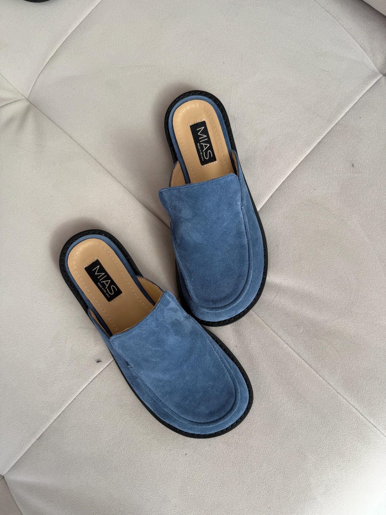 May include: A pair of blue suede slip-on shoes with black soles and the word "MIAS" printed on the inside of each shoe.