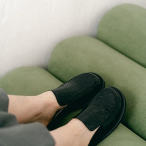 May include: A pair of black leather slip-on shoes with a crocodile-embossed pattern. The shoes are worn by a person sitting on a green sofa.