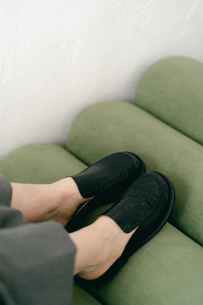 May include: A pair of black leather slip-on shoes with a crocodile-embossed pattern. The shoes are worn by a person sitting on a green sofa.